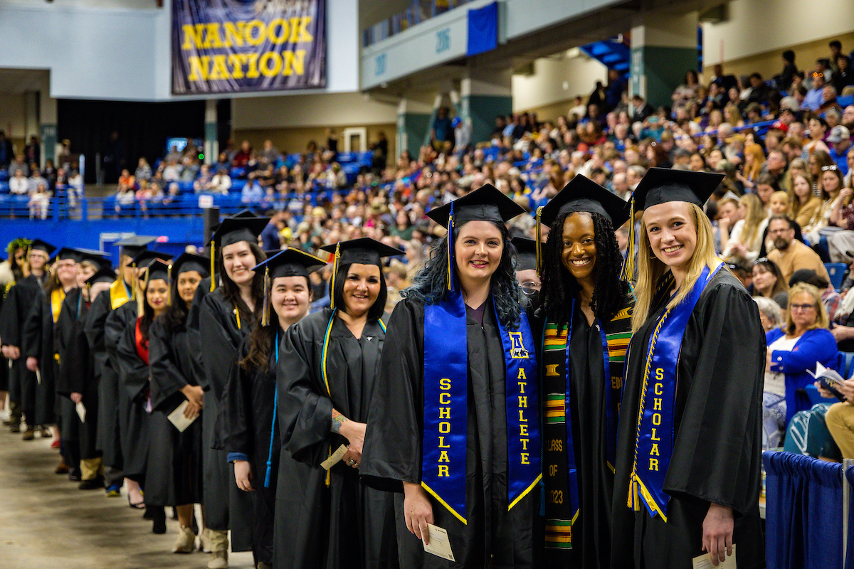 A line of college graduates wearing mortarboards and black academic robes and regalia standing inside an arena. A sign in the background reads, “Nanook Nation.”